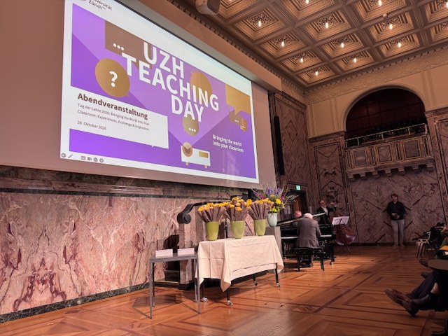 A grand hall with a large screen showing the UZH Teaching Day 2026 evening event slide, flowers on a table in front, and musicians at a piano.
